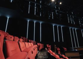 Rij roodpluche theaterstoelen in een donkere zaal met zwarte wanden en verticale LED-lijnverlichting; links de tribune, rechts de rand van het podium/techniekruimte.