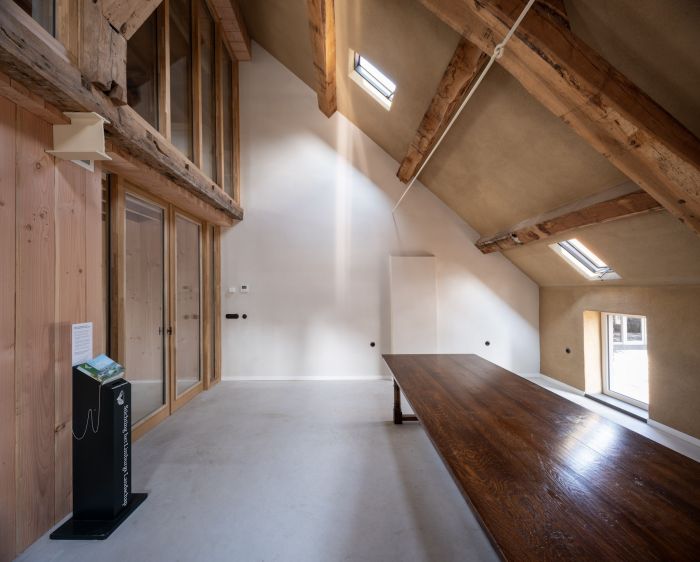 Zolderruimte van de Grathemermolen met zichtbare houten spanten, hoge schuine plafonds met dakramen, glazen scheidingswand links en een lange houten tafel langs het raam.