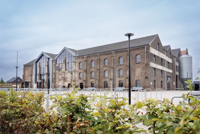 Getransformeerde Greswarenfabriek in Reuver: gerestaureerd bakstenen fabriekspand met grote glazen entree, oude gevelletters en silo aan het plein met fietsenrekken en lantaarnpalen.