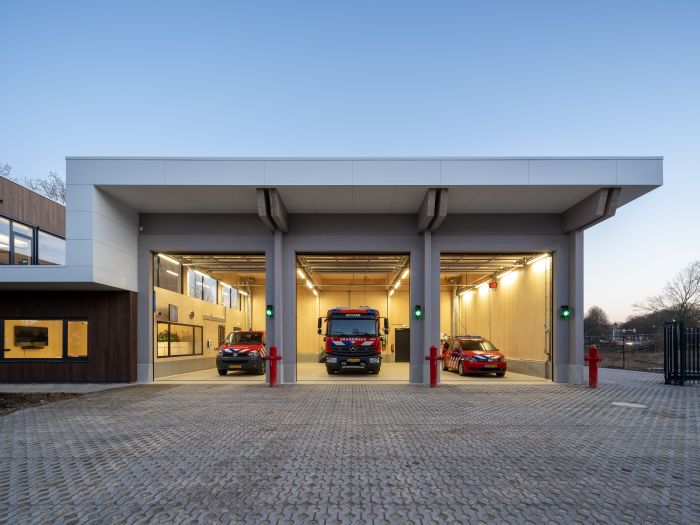 Front van de nieuwe brandweerpost Sittard-Oost in de schemering met drie open uitrijpoorten; links een brandweerbus, midden een tankautospuit en rechts een dienstauto, allemaal klaar voor uitruk op het verlichte terrein.