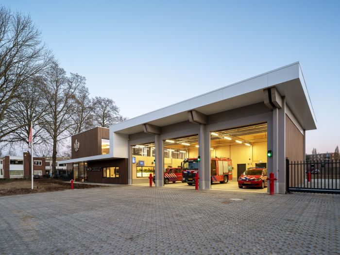 Nieuwe brandweerpost Sittard-Oost bij schemering met drie open uitrijpoorten; binnen staan een brandweerwagen en dienstauto klaar, naast de glazen entree in het moderne hout-wit gebouw.