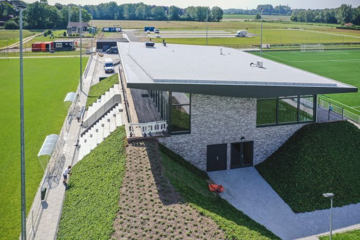 Schuin luchtperspectief van het clubgebouw van DVO’20 met groot uitkragend dak, bakstenen gevel en glazen pui, ingebouwde traptribune tussen groene taluds en dug-outs langs het hoofdveld.