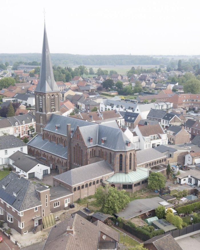 Luchtfoto bakstenen kerk met hoge torenspits, kloostergang en zinken daken midden in dorpsker