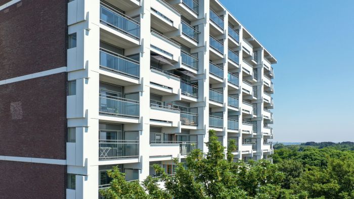 Close-up van de gerenoveerde galerijflat met witte gevelelementen en glazen balkonhekken; bakstenen kopgevel links, bomen en blauwe lucht op de achtergrond.