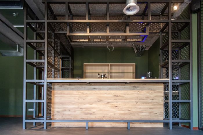 Interieur MFA Treebeek – industriële bar/balie met houten front, zwart stalen rekwerk en zichtbare installaties aan plafond