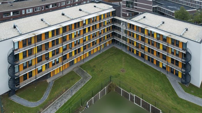 Nieuwbouw met twee vleugels in een L-vorm; galerijen met gele en oranje panelen, metalen balustrades en buitentrappen rond een grasrijke binnentuin.
