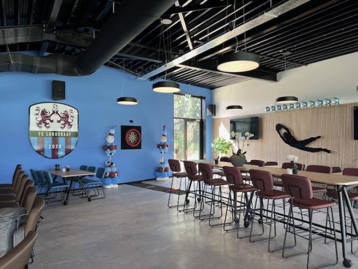 Interieur clubgebouw FC Landgraaf: blauwe wand met clublogo, hoge tafels, barstoelen en sportdecor