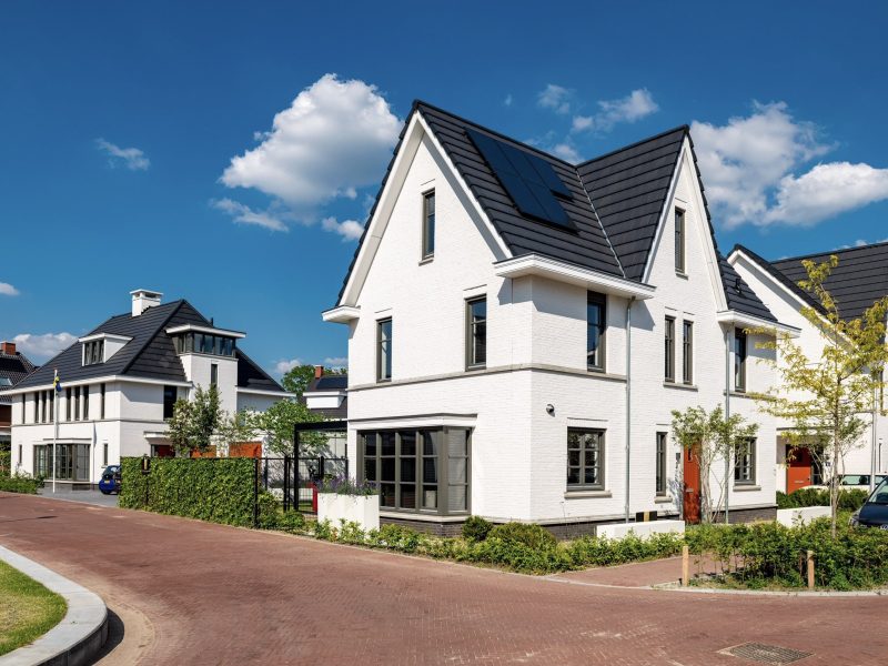 Moderne vrijstaande witte woning met donker zadeldak en zonnepanelen in een rustige woonwijk op een zonnige dag.