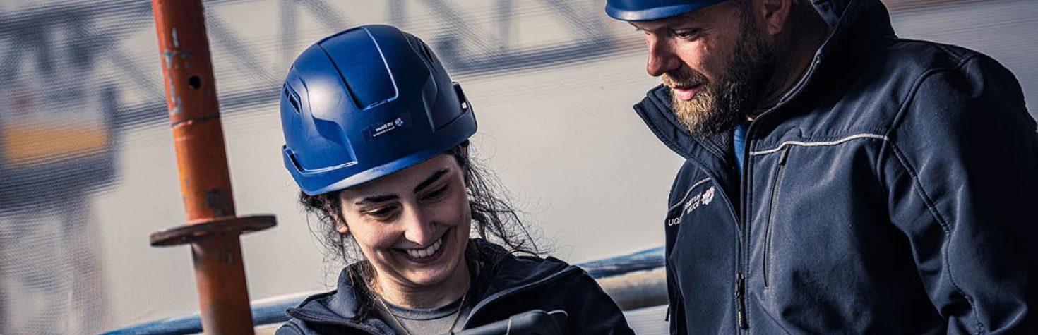 Twee mensen met blauwe bouwhelmen kijken lachend naar een tablet op een bouwlocatie.