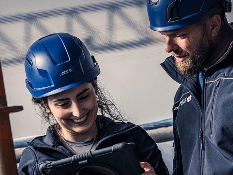 Twee mensen met blauwe bouwhelmen kijken lachend naar een tablet op een bouwlocatie.